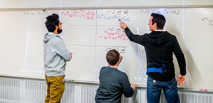 Professor Matt Salomone points to part of an equation written on a white board by a student who stands with a marker in his hand listening to the professor while another student works on an equation on the board