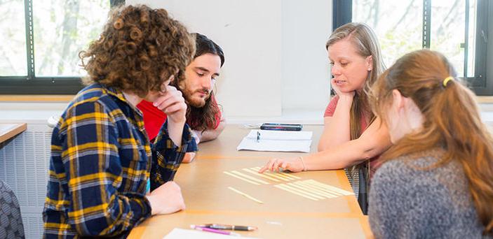 Professor Shannon Lockard sits at a classroom table with 3 students explaining something with strips of paper laid out on the table