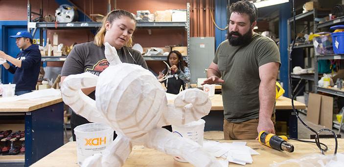 BSU student and woodworking professor discuss the student's project, a wooden framed octopus covered in white fabric, while 2 other students work in the background