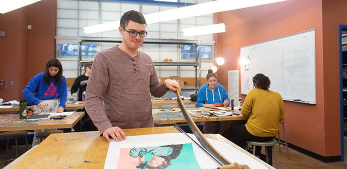a student trims his artwork with a large paper cutter while other students work on their pieces at tables behind him